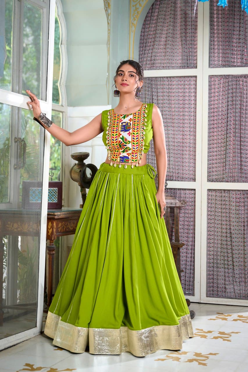 Woman in a green traditional outfit with intricate patterns standing indoors.